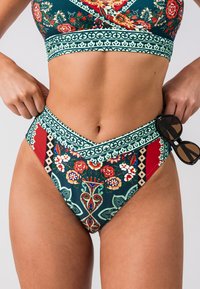 Completo bikini a due pezzi con motivo floreale e geometrico in tonalità di verde, rosso e crema. Design a vita alta, completato da occhiali da sole.