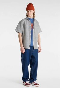 Kortærmet stribet skjorte i sort og hvid over en blå grafisk t-shirt, kombineret med løstsiddende blå jeans og røde sneakers, toppet med en orange hue.