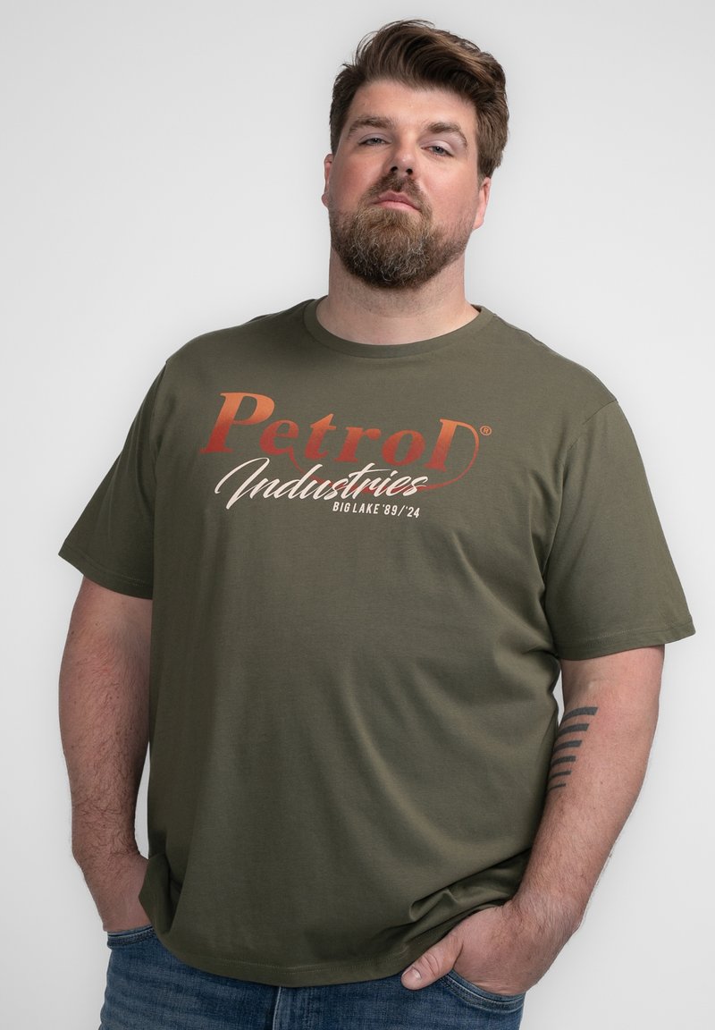 Camiseta verde oliva de corte relajado, con un texto en negritas y multicolor que dice "Petrod Industries" y "Bio Lake '89/24" en un diseño semicasual.