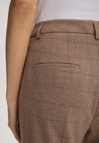 Bruine houndstooth broek met rood ruitpatroon, heeft een enkele achterzak en een platte tailleband. Materiaal lijkt textuur te hebben.