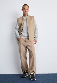 Hombre de pie contra una pared blanca, vistiendo un chaleco beige, un suéter gris de canalé, pantalones beige holgados y zapatillas negras y amarillas.