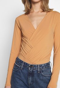 Langärmliger Body in Beige mit tiefem V-Ausschnitt, mit gewickeltem Vorderdesign und gerafftem Taillenbereich. Kombiniert mit hoch taillierten Jeans.
