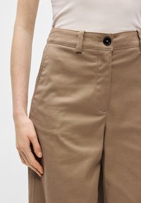 Gai brūni bikses, kas izgatavotas no teksturētas auduma. Ar jostu cilpiņām, pogu aizdari un sānu kabatām. Pamanāmi šuvju detaļas.