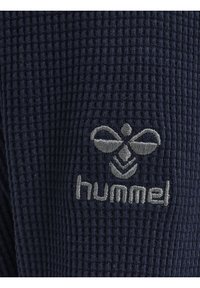 Navyblaue, waffelstrukturierte Stoff mit einem grauen, gestickten Logo, das ein Bienenmuster zeigt, und dem Text "hummel" unterhalb des Logos.