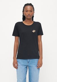 Svart bomullst-skjorte med korte ermer, rund halslinning, og et hjerte-design av gullpaljetter på venstre bryst. Brukt med lyseblå jeans.