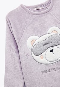 Sudadera de forro polar morada con diseño de cara de oso, que incluye una máscara de dormir bordada y texto, textura suave, escote redondo y mangas raglán.