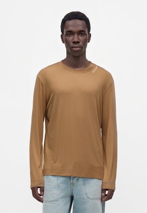 Ung mand med kort hår iført en ensfarvet brun langærmet skjorte og lyseblå jeans, stående mod en hvid baggrund.
