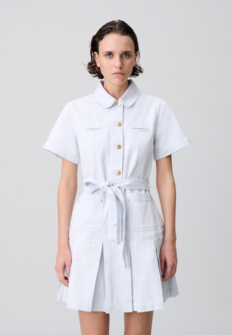 Robe chemise en denim bleu clair avec des manches courtes, un devant boutonné, une ceinture attachée et deux poches avant. Présente un détail de jupe plissée.