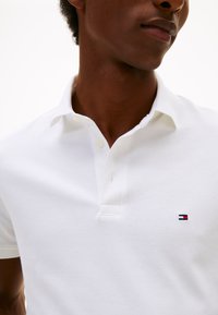 Hvit poloshirt laget av teksturert bomullsstoff, med to-knaps knepping og en liten logo på venstre bryst i blått, rødt og hvitt.