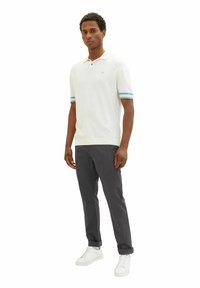 Weißes Poloshirt mit kurzärmligen blauen Streifen, kombiniert mit dunkelgrauen Hosen und weißen Sneakern. Einfaches Design und schmaler Schnitt.