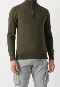 Olivgrüner Pullover mit hohem Kragen, Front mit halbem Reißverschluss und gerippten Bündchen. Dazu hellgraue Cargohosen mit Seitentaschen.