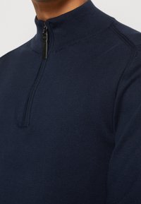 Primo piano di una persona che indossa un maglione a maglia blu navy con collo a costine a girocollo e una linguetta di cerniera nera.