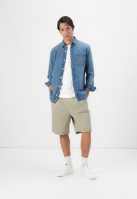 Denim overhemd met knopen in het blauw, een witte t-shirt eronder, kaki shorts, witte sneakers, staand met de handen in de zakken, neutrale achtergrond.
