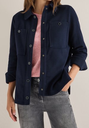 Frau trägt eine marineblaue Jacke mit Knöpfen über einem rosa Top und grauen Jeans, mit einer Hand in der Tasche und hochgekrempelten Ärmeln.