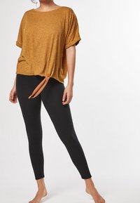 Sennepsgul kortærmet top med knyttet detalje i taljen, parret med sorte leggings; blød tekstur og afslappet pasform.