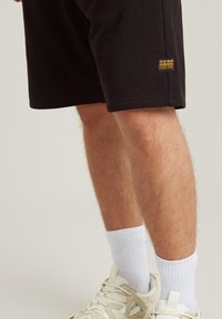Bermuda nera in cotone con un piccolo logo G-Star. Modello che indossa calzini bianchi e sneakers color crema, mostrando un outfit casual.
