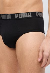 Ανδρικά μαύρα εσώρουχα τύπου briefs με φαρδιά μέση που φέρει το λογότυπο "PUMA", φορεμένα από ένα άτομο που στέκεται, από τη μέση έως το μέσο του μηρού, με το δεξί χέρι ορατό.