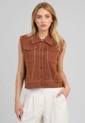Femme blonde portant un gilet marron sans manches boutonné avec des surpiqûres blanches et un pantalon plissé blanc taille haute, main dans la poche, fond neutre.
