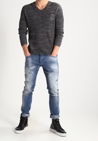 Ciemnoszary, teksturowany sweter z dzianiny z dekoltem w serek i małą kieszonką, zestawiony z jasnoniebieskimi, postarzanymi dżinsami i czarnymi wysokimi trampkami.