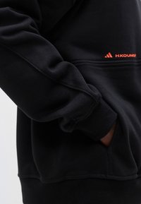 Fekete pulóverujj és kéz az oldalzsebben, apró narancssárga Adidas logóval és "H.KOUMORI" felirattal az elején.