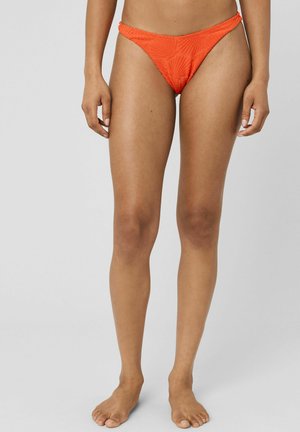 Femme portant un bas de bikini orange texturé, debout pieds nus sur un fond clair uni.