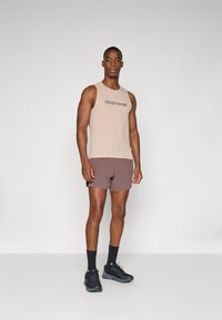 Beige ärmelloses Tanktop mit schwarzem Logo, kombiniert mit braunen Sportsch shorts. Modell trägt schwarze Sneakers und Socken in Crew-Länge.