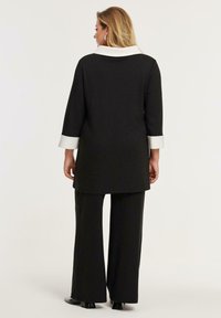 Frau, die mit dem Rücken zur Kamera steht, trägt schwarze weit geschnittene Hosen und ein schwarzes Langarmoberteil mit weißen Akzenten an Kragen und Manschetten, vor einem schlichten Hintergrund.