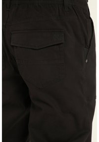 Pantaloni cargo neri con un design aderente, numerose tasche e un tessuto testurizzato. I dettagli degni di nota includono bottoni metallici e un logo ricamato.