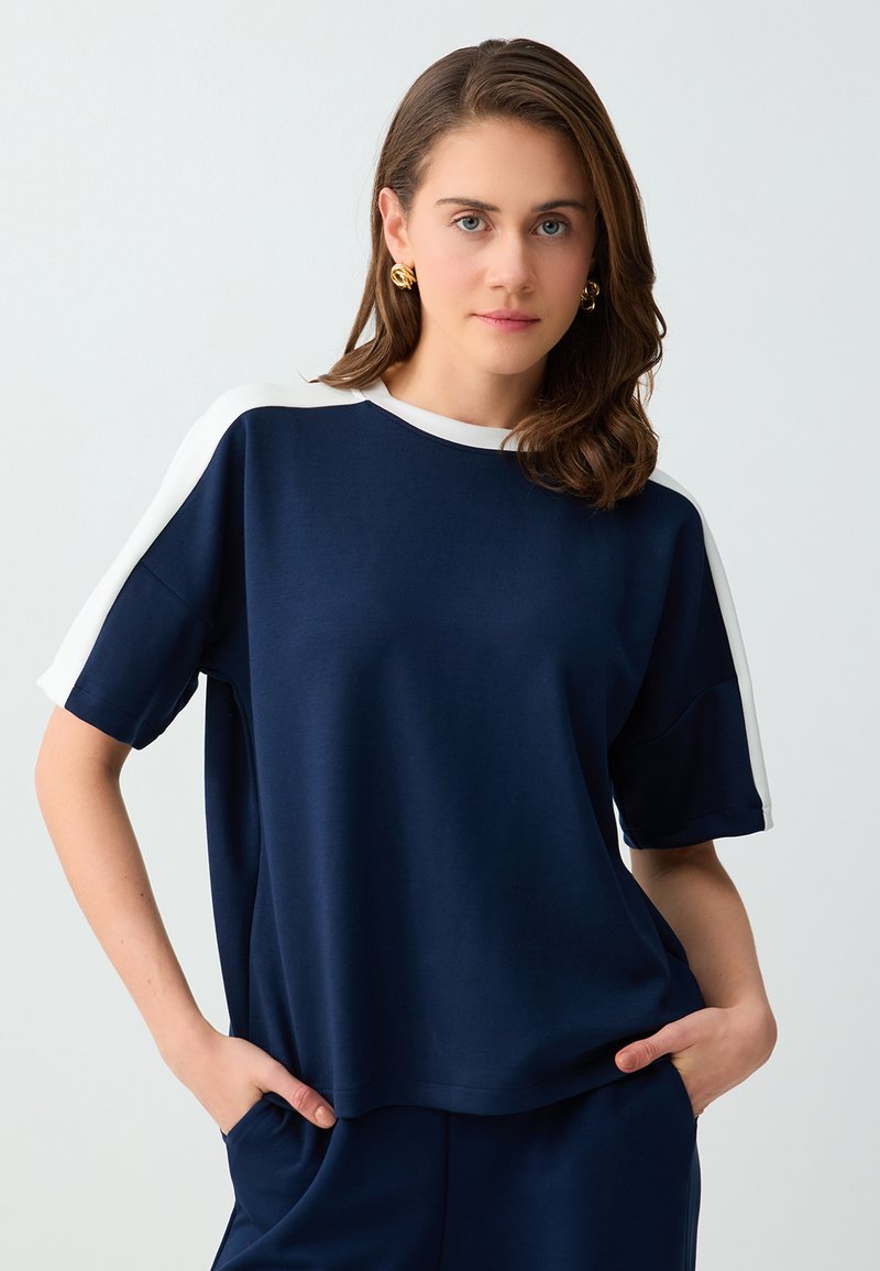 Femme aux cheveux bruns portant un haut bleu marine à manches courtes avec des bandes blanches aux épaules et des boucles d’oreilles créoles dorées, mains dans les poches, fond neutre.