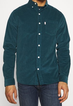 Uomo che indossa una camicia di velluto a coste teal con bottoni bianchi e una tasca frontale sul petto, abbinata a jeans blu scuro.