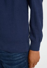 Marineblå teksturert genser med ribbestrikket kant og mansjetter, med en sidemerke detalj, vist på en hånd med mørke jeans.