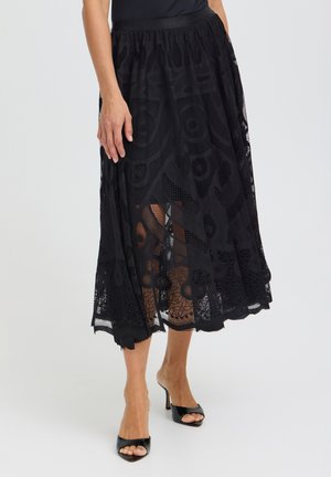 Femme portant une jupe midi en dentelle noire à motifs, un haut noir et des chaussures à talons hauts noires ouvertes à l'avant, sur fond blanc.