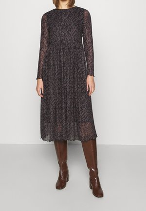 Femme portant une robe mi-longue noire à manches longues avec un petit imprimé floral et des bottes marron montantes jusqu'aux genoux, se tenant devant un fond blanc uni.