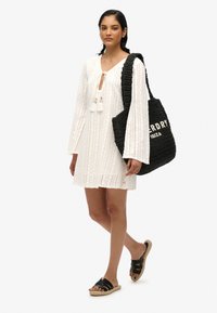 Witte, lange mouwen jurk met een strik halslijn en franjes. Geweven zwarte tote bag met de tekst "ERDRY IBIZA". Zwarte schuif sandaal met banden.