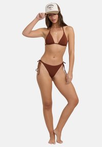 Biquíni castanho com top em forma de triângulo e cuecas com laço nas laterais. Combinado com um boné de camionista bege com um design gráfico. Textura suave e lisa.