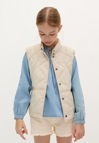 Gilet matelassé beige sur une chemise bleu clair à manches bouffantes, avec fermeture à boutons et poches latérales. Shorts beiges frangés courts.
