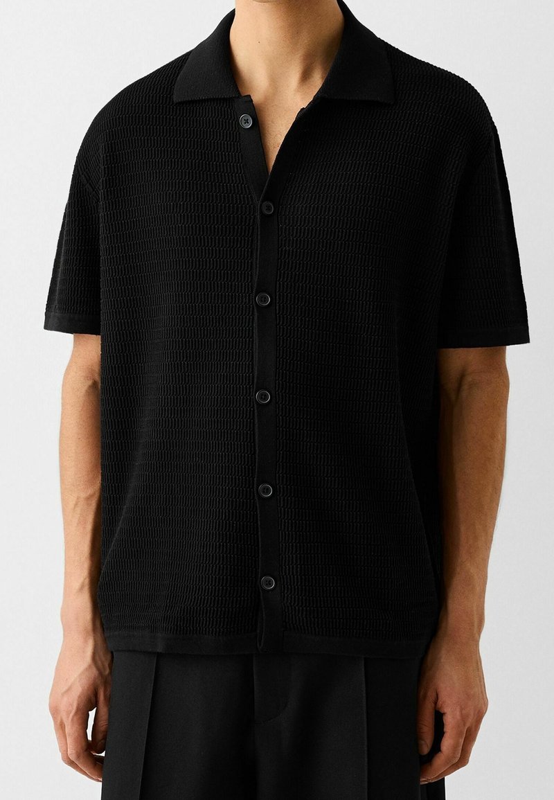 Polo shirt nero a maniche corte realizzato in tessuto testurizzato con una trama decorativa, dotato di chiusura con bottoni e colletto classico.