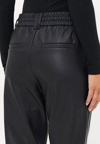 Zwarte nep leren broek met een elastische tailleband, twee achterzakken en een gladde textuur, getoond vanuit de zij- en achteraanzicht.