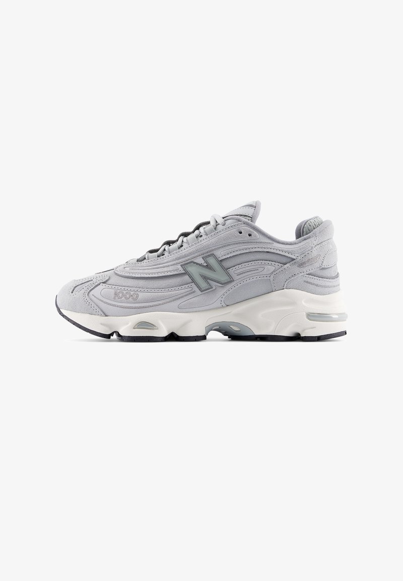 Γκρι αθλητικό παπούτσι με επάνω μέρος από σουέντ και δίχτυ, σχεδιασμό με υφή, μαύρη σόλα, αντανακλαστικές λεπτομέρειες και εμφανές λογότυπο "N" στο πλάι.