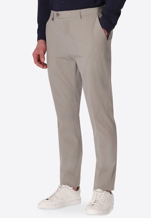 Uomo che indossa pantaloni beige slim fit con tasche laterali e sneaker bianche su sfondo semplice.