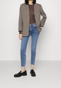 Žena v taupe saku, průhledném hnědém topu, modrých džínách a černých mokasínech, stojící s jednou rukou v kapse na bílém pozadí.