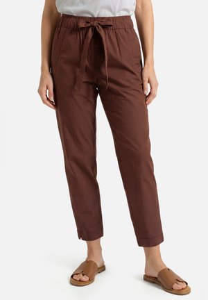 Person wearing high-waist, brown tapered pants with a drawstring bow and slip-on tan sandals on a white background.