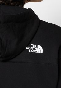 Gros plan d'une personne portant un sweat à capuche noir avec le logo blanc "The North Face" sur le haut du dos, à droite.