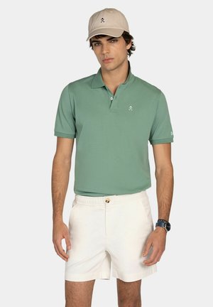 Giovane uomo che indossa un berretto beige, una polo verde a maniche corte, pantaloncini bianchi e un orologio da polso scuro, in piedi davanti a uno sfondo semplice.