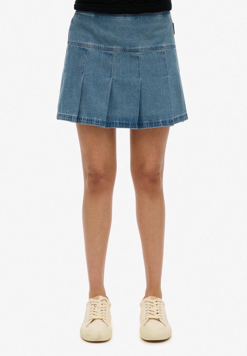 Falda de mezclilla plisada en azul claro, con una cintura ajustada y un dobladillo acampanado. Combinada con zapatillas beige con cordones para un look casual.