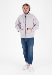 Lichtgrijze hoodie met een ritssluiting aan de voorkant, grote zakken en een capuchon met trekkoord, gecombineerd met blauwe jeans en witte sneakers.