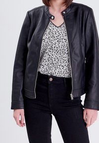 Veste en cuir noir avec un col rond, une fermeture éclair et une texture lisse, portée sur un haut blanc à motif floral noir.