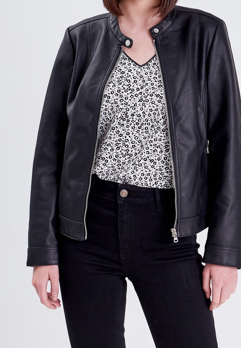 Veste en cuir noir avec un col rond, une fermeture éclair et une texture lisse, portée sur un haut blanc à motif floral noir.