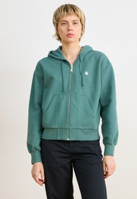 Grønn zip-up hettegenser laget av mykt stoff med kengurulomme, snorer på hetten og en liten logo på brystet.