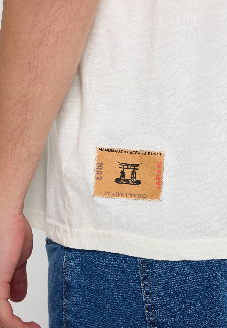 Kremowa koszulka z krótkim rękawem z teksturowanej tkaniny. Posiada beżową metkę wszytą na dolnym końcu, oznaczoną napisem "HANDMADE BY SASAKIJIRUSHI" i "JAPAN."